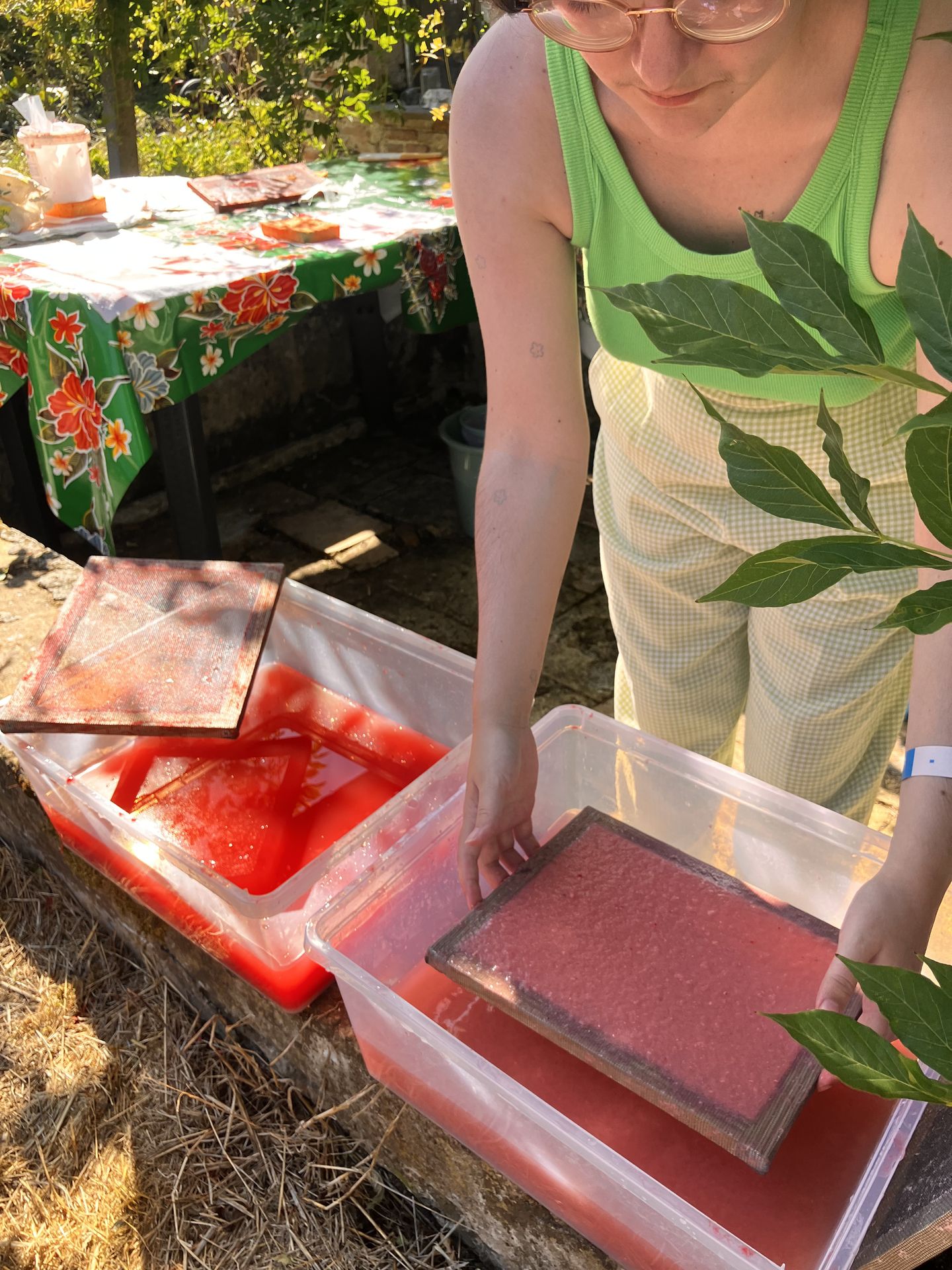 Les mains de solaeflore en train de fabriquer du papier en extérieur. Il tient la forme au dessus d'un bac d'eau rempli de pulpe rose.